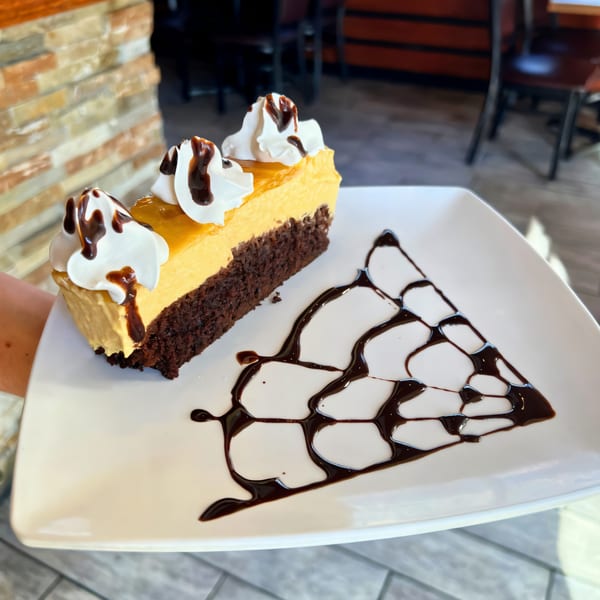 Slice of Peruvian lucuma cheesecake with chocolate drizzle and whipped cream on a white plate.