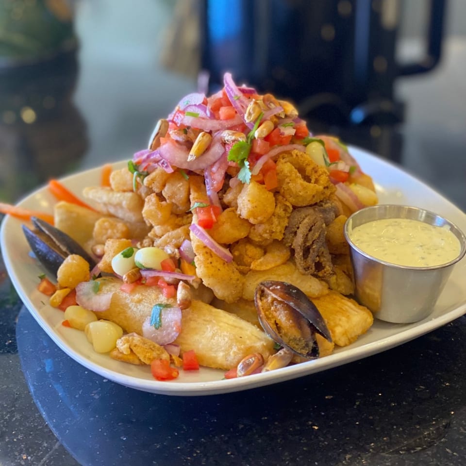 A large platter of Peruvian Jalea Mixta with fried seafood, mussels, red onions, and a side of green sauce.