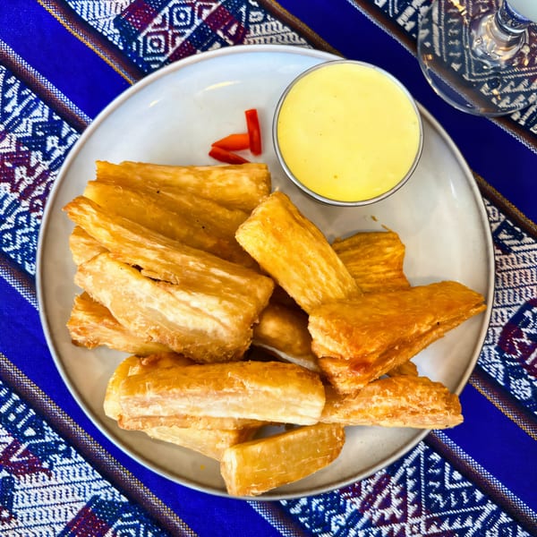 Golden fried yuca sticks with a side of yellow dipping sauce and red pepper slices on a patterned tablecloth.
