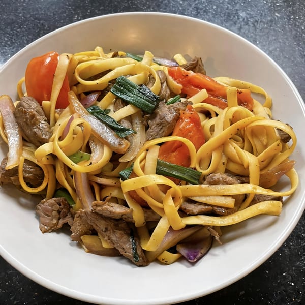 Peruvian Lomo Saltado with fettuccine, beef, tomatoes, and green onions in a white bowl.